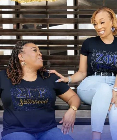 Two women sitting together, wearing matching black t-shirts adorned with "ΣΙΠ Sisterhood" and "ΖΦΒ" in sparkling letters, showcasing camaraderie and sisterhood.