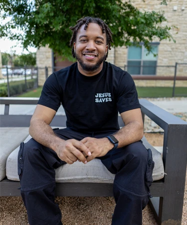 Man sitting on a bench wearing a black "Jesus Saves" t-shirt, smiling, with greenery and a building in the background, representing faith-based clothing for Walk In Faith Clothing.