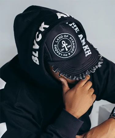 Person wearing a black hoodie and a distressed cap with "BLVCK ANKH" and "LAND OF PYRAMIDS" text, showcasing urban fashion style relevant to The Print Lounge by Blvck Ankh.