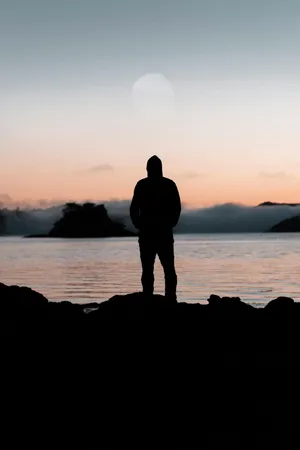 Silhouette of a person in a hooded jacket standing on rocks by a serene body of water at sunset, symbolizing creativity and reflection in a tranquil setting.