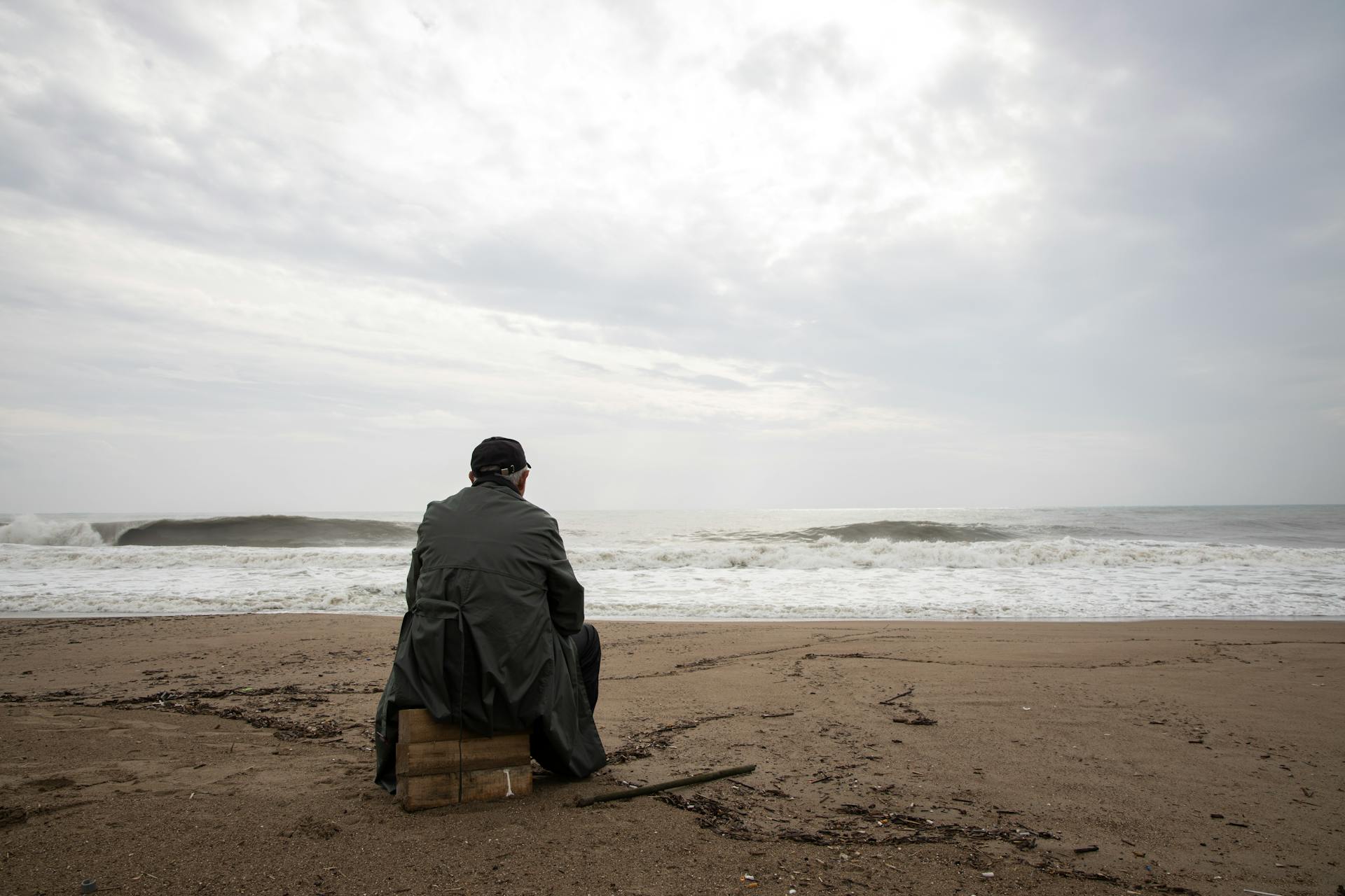 Person sitting on a wooden crate at the beach, gazing at the ocean waves under a cloudy sky, reflecting solitude and contemplation, symbolizing creativity and collaboration in overcoming challenges.