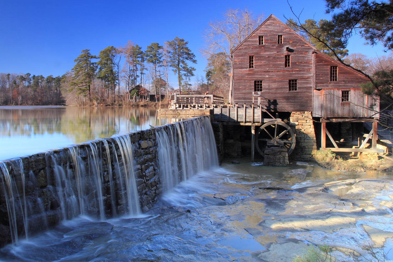 Historic watermill beside a serene lake, featuring a wooden structure with a waterwheel and cascading waterfall, surrounded by trees and a clear blue sky, symbolizing Raleigh's local heritage and natural beauty.