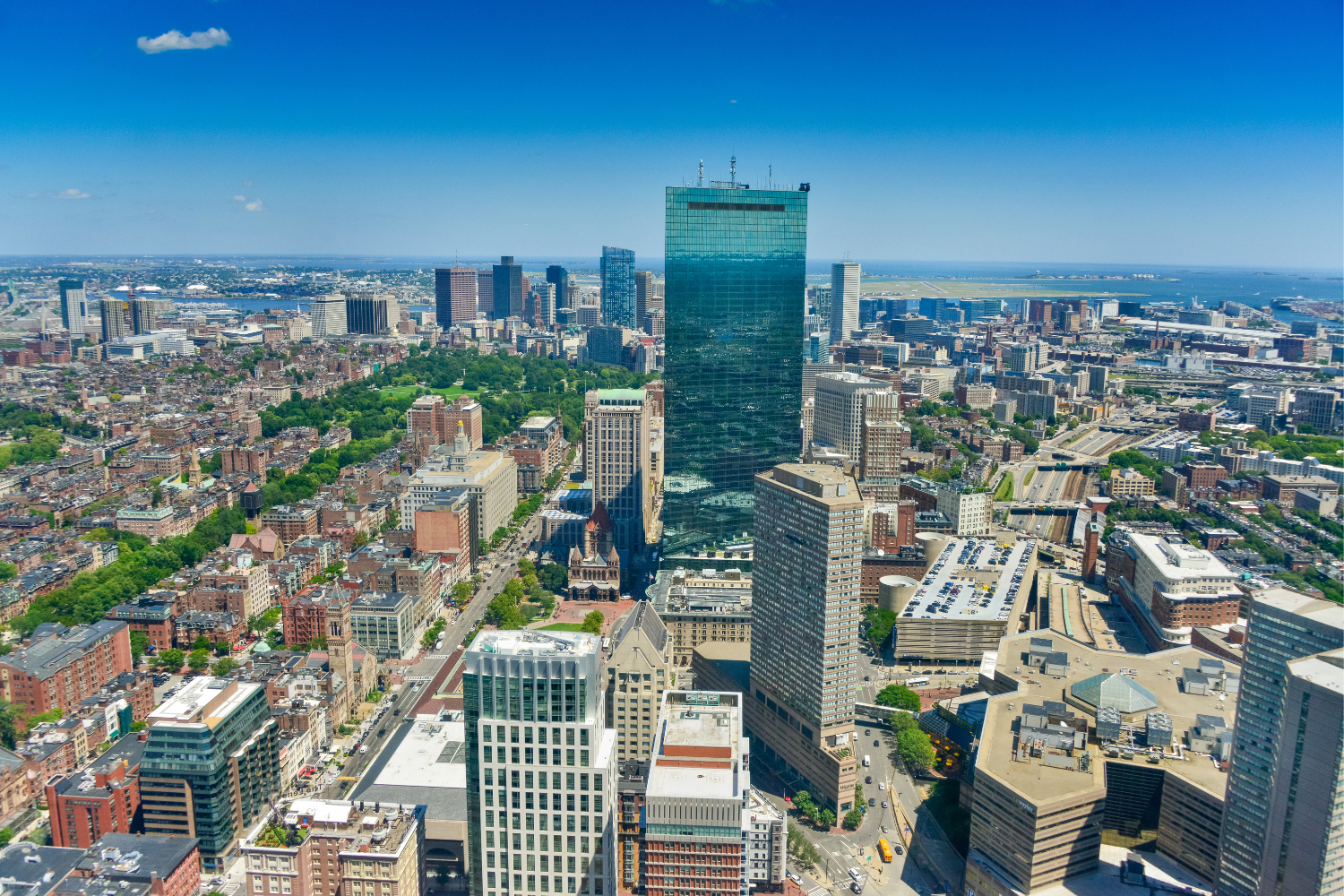 Aerial view of Boston's skyline featuring the tall glass structure of the John Hancock Tower, surrounded by urban buildings and greenery, illustrating the city's modern architecture and layout.