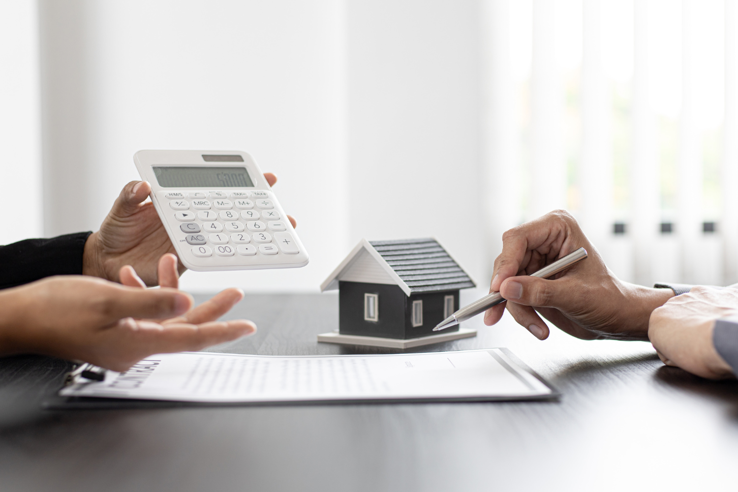Hands holding a calculator and a pen, discussing a model house on a table, symbolizing financial planning and decision-making for roofing and fulfillment strategies.