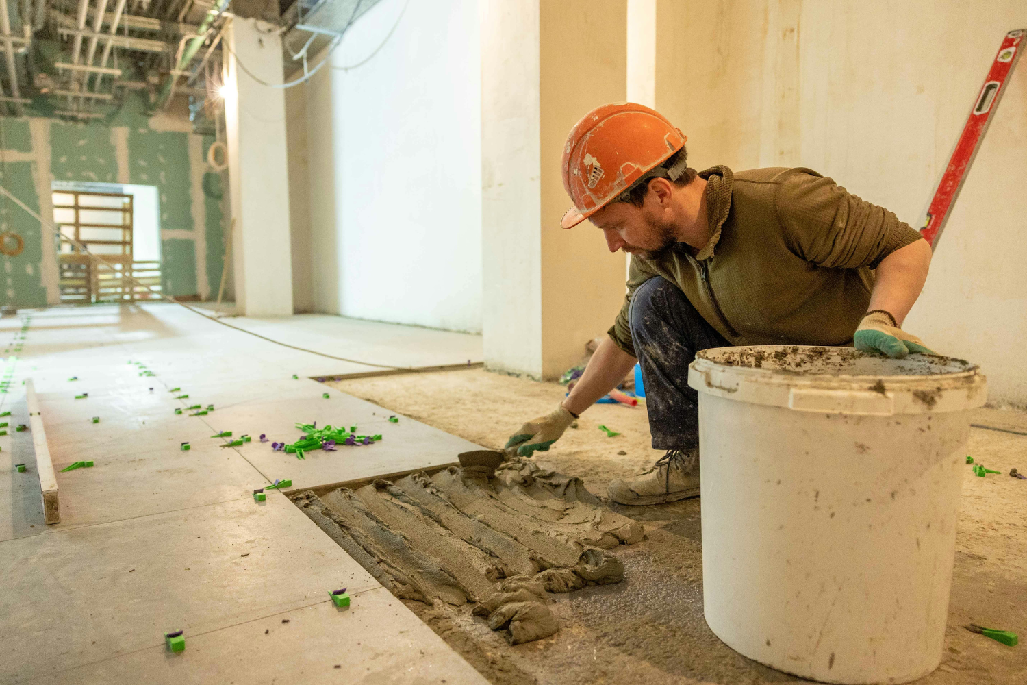 Construction worker laying grout on a floor, wearing an orange hard hat and gloves, in a partially finished interior space with scattered tile spacers.