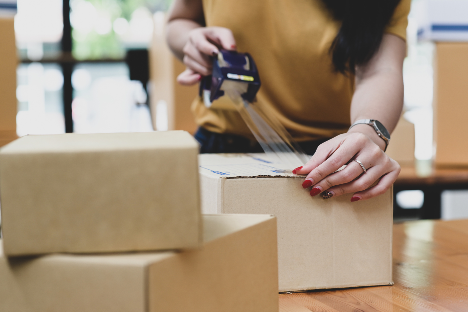 Woman sealing a cardboard box with packing tape in a workspace, emphasizing eCommerce logistics and shipping preparation.