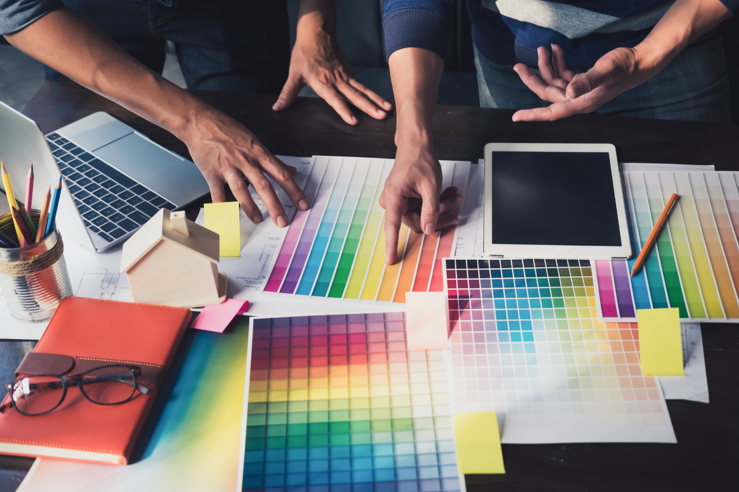 Hands discussing color palette options over design materials, laptop, and tablet on a workspace, highlighting brand identity development for marketing strategies.