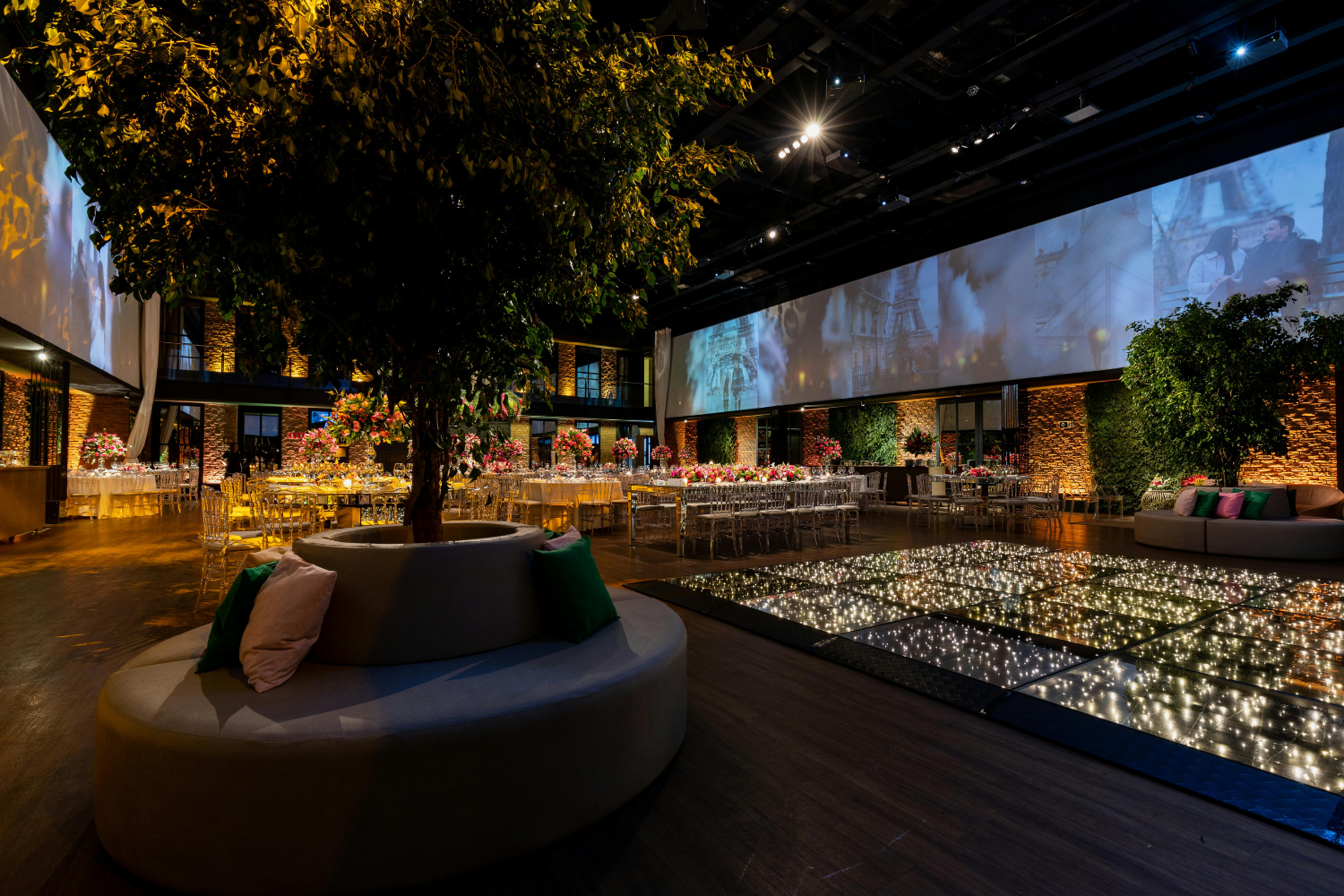 Elegant event space featuring a central decorative tree, surrounded by tables set for dining with floral arrangements, illuminated by ambient lighting and a display screen showcasing visuals, emphasizing a sophisticated atmosphere for branding events.