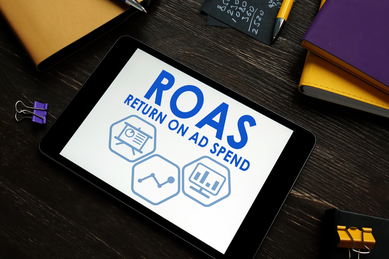 Tablet displaying "ROAS" and "RETURN ON AD SPEND" with graphical elements, surrounded by office supplies, emphasizing PPC marketing effectiveness and ad spend optimization.