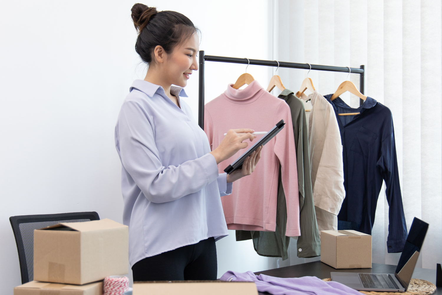 Woman using a tablet to manage email marketing for an online clothing store, surrounded by clothing items on hangers and shipping boxes, illustrating e-commerce engagement strategies.