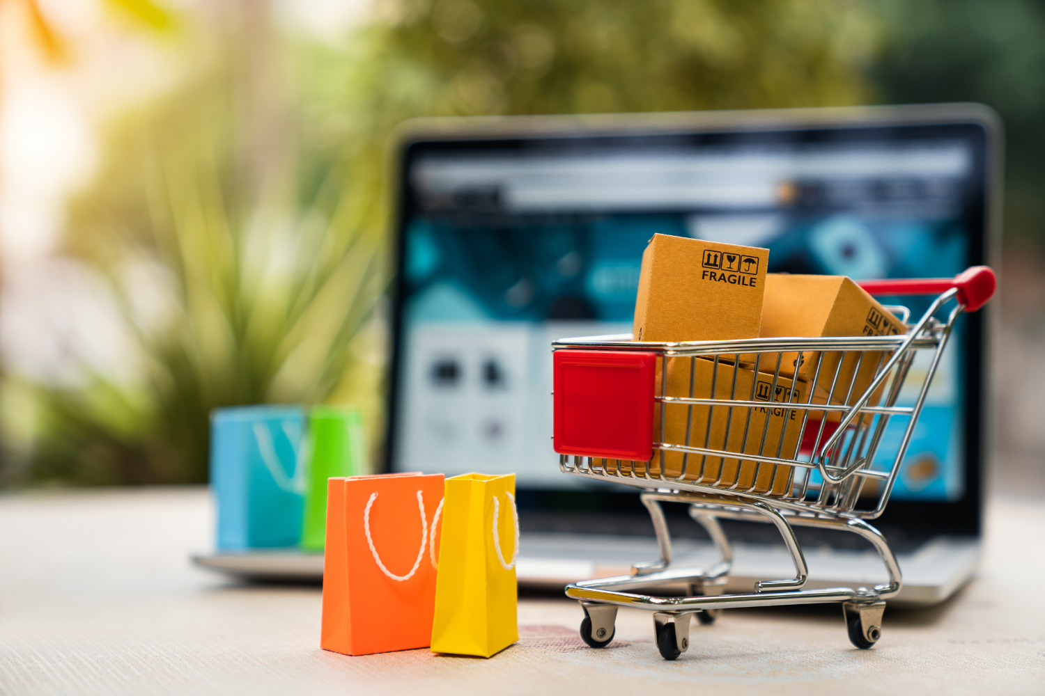 Shopping cart with fragile packages in front of a laptop screen, colorful shopping bags nearby, representing e-commerce and online shopping strategies for effective PPC campaigns.
