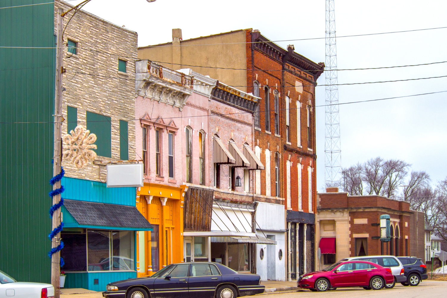 Colorful storefronts along a street featuring a mix of architectural styles, including brick and stone, with parked cars and a radio tower in the background, emphasizing local business presence and community engagement.