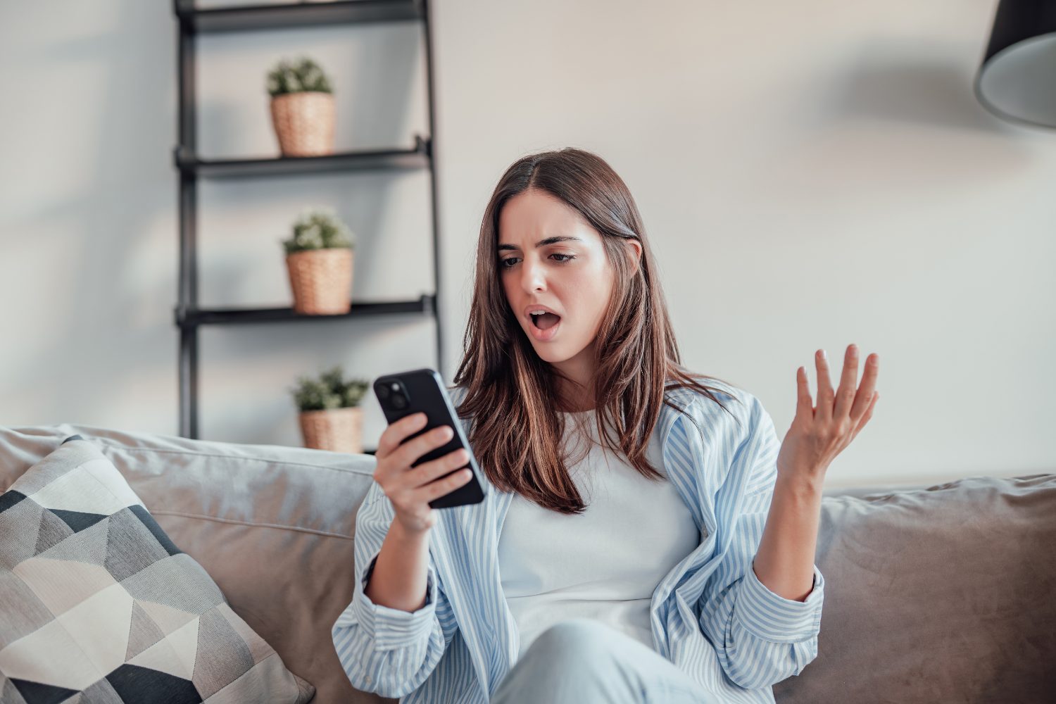 Woman expressing frustration while looking at smartphone, sitting on couch in a modern living room, with decorative plants in the background, relevant to digital marketing insights.