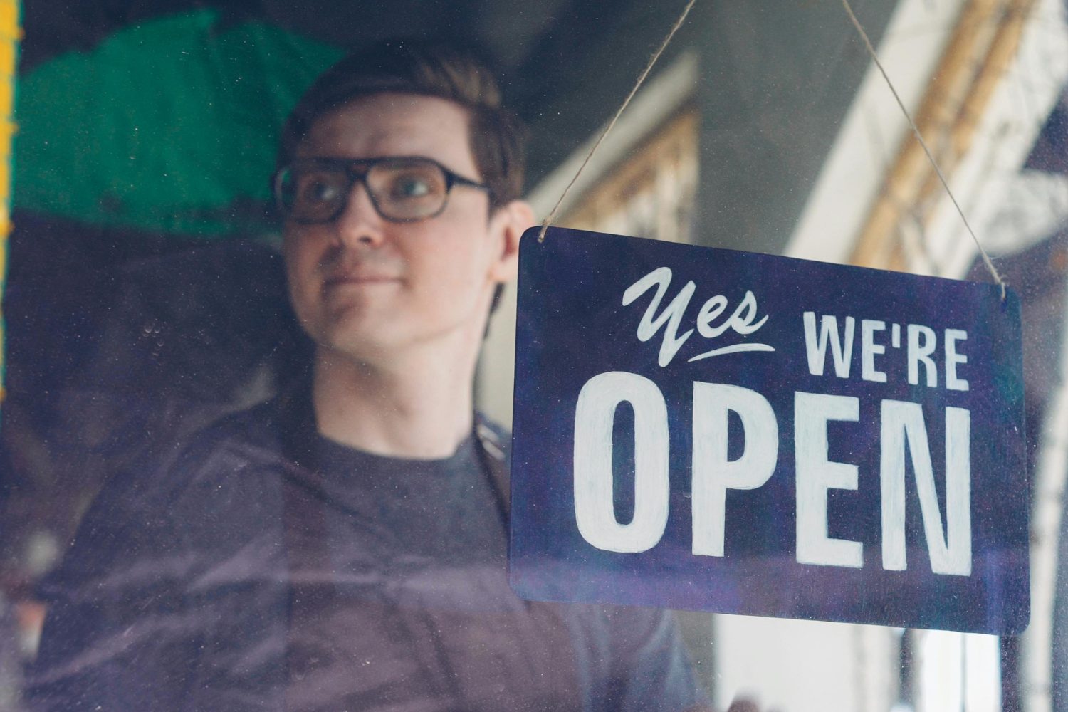 Man smiling behind a "Yes, We're Open" sign, symbolizing small business accessibility and customer engagement in the context of effective advertising strategies.