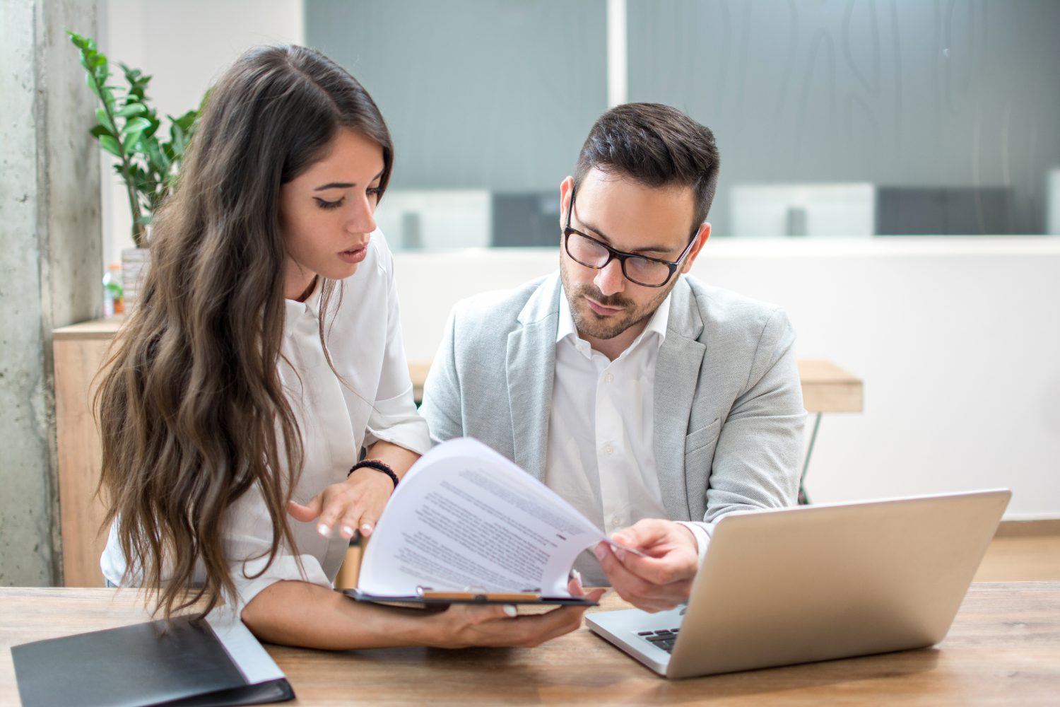 Two professionals discussing digital marketing strategies, reviewing documents, with a laptop on the table, in a modern office setting.